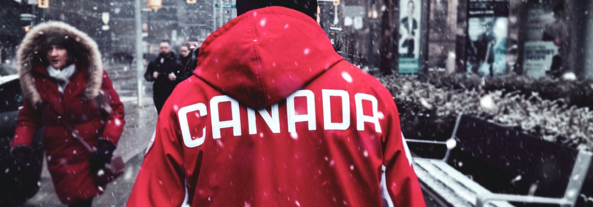 A man wearing a red jacket with 'Canada' written on the back walks down a city street as it snows.