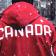 A man wearing a red jacket with 'Canada' written on the back walks down a city street as it snows.