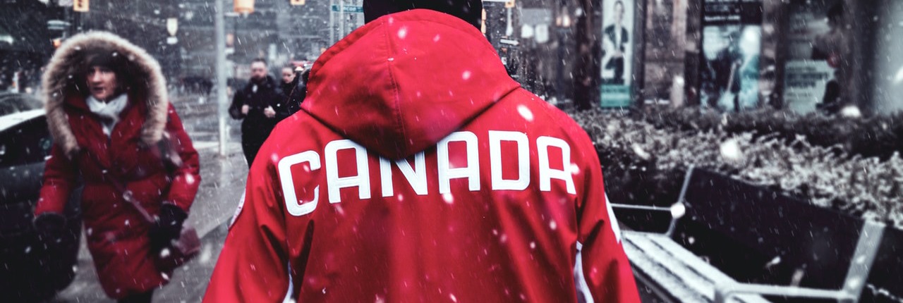 A man wearing a red jacket with 'Canada' written on the back walks down a city street as it snows.