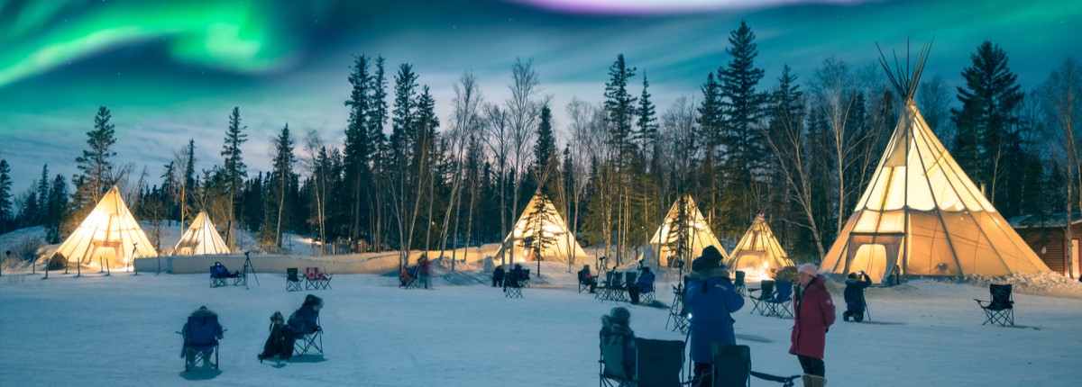 People in winter coats stand near illuminated tents and camping chairs in a wintry scene