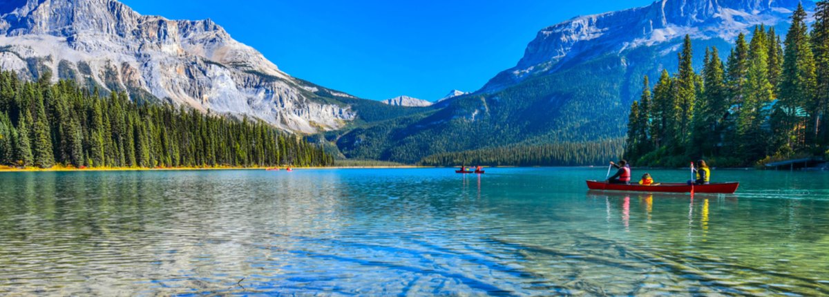 Kayakers paddle across a lake bordered by forest, with mountains in the background