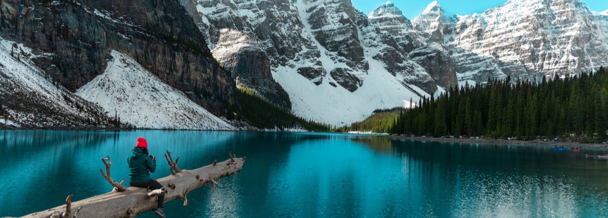 A log stretches out above a lake in front of steep, snowy mountains. A woman is sitting on the log taking a photograph with a camera.