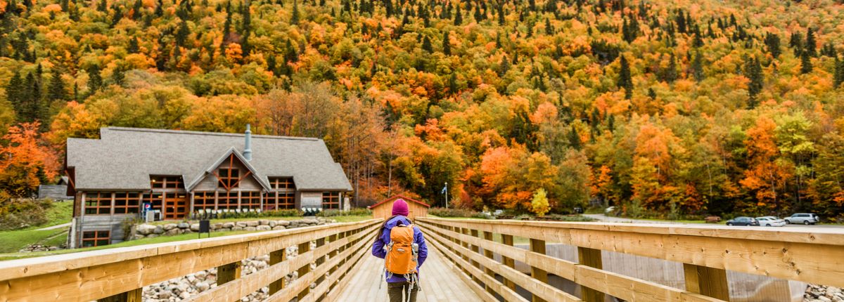 A backpacker walks across a wooden bridge toward forested hills rick in autumn colours