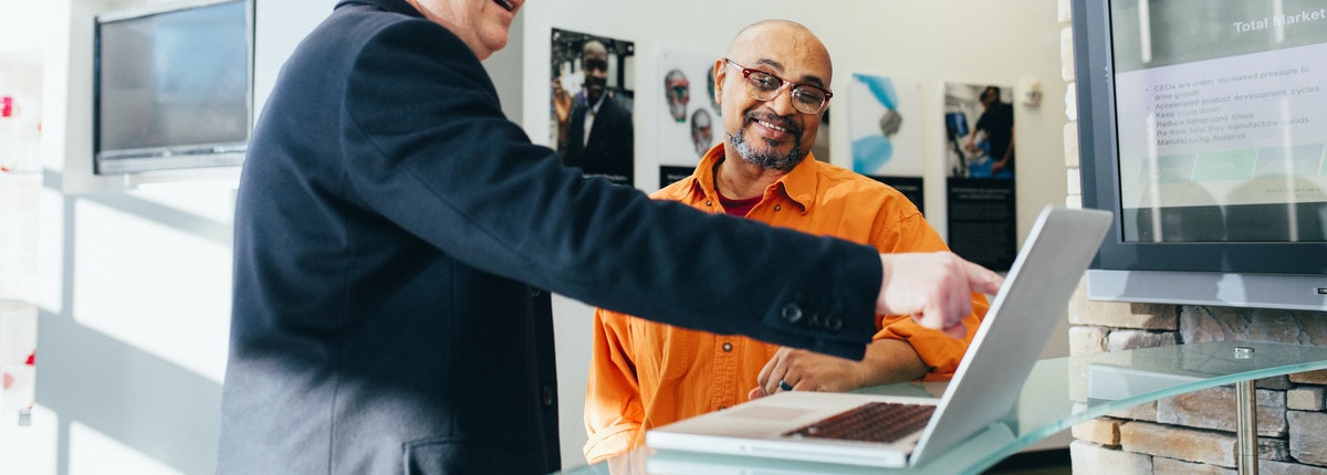 A man in a suit points at a laptop screen while a second man in a shirt looks on